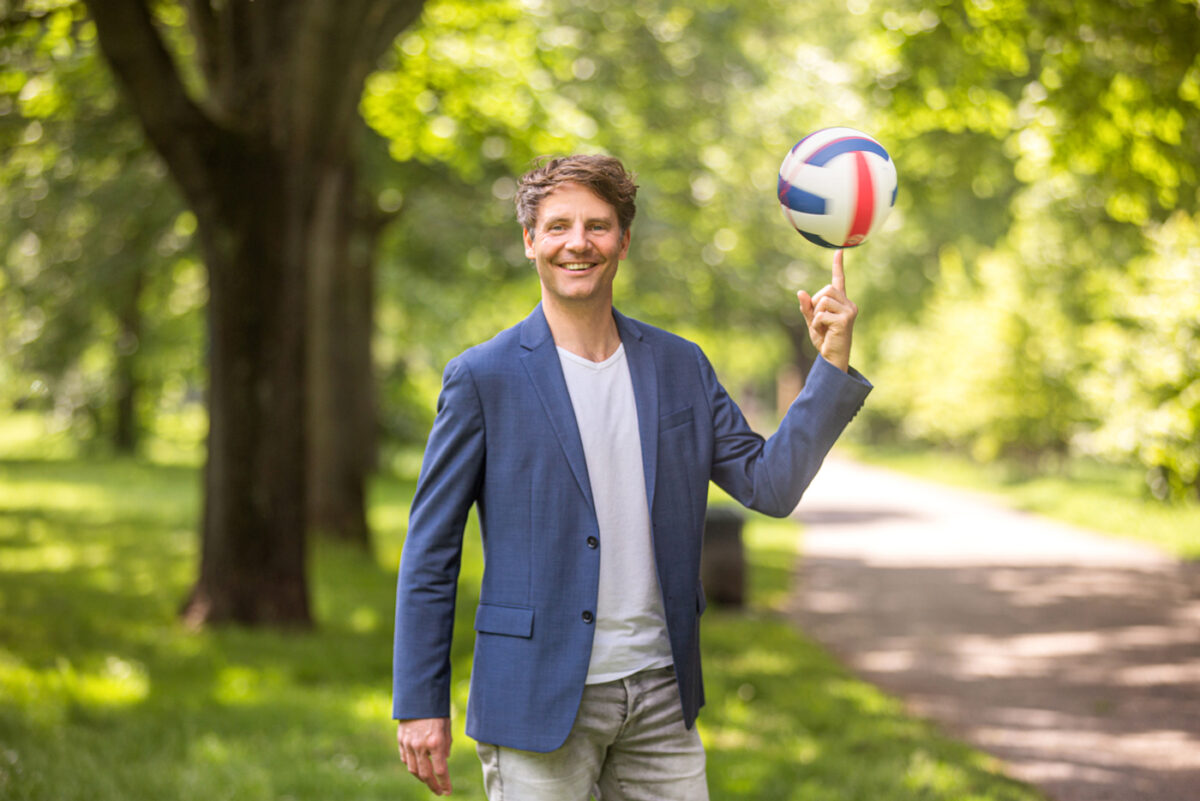 Personal Branding Foto vom Sportcoach in natürlichem outdoor Setting. Der Coach guckt freundlich in die Kamera. Fotografiert er einen Volleyball auf seinem Finger rotiert.